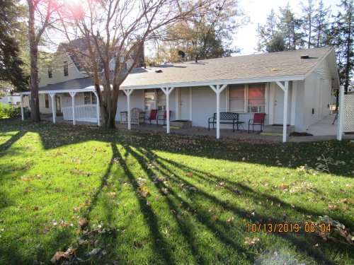 A white house with a porch, surrounded by green grass and trees, casting long shadows on a sunny day.