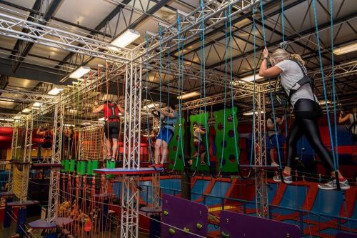A group of people navigating an indoor ropes course with colorful obstacles and safety harnesses.