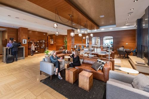 A modern hotel lobby with a cozy seating area, wooden accents, and guests chatting near a reception desk.