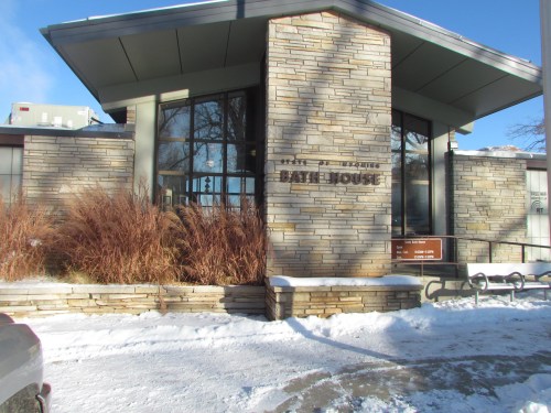 A modern stone building with large windows, labeled "Bath House," surrounded by snow and winter vegetation.