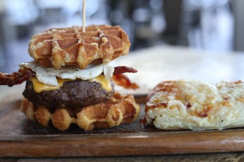 A stacked burger with waffles, bacon, and an egg, served alongside a cheesy potato dish on a wooden platter.