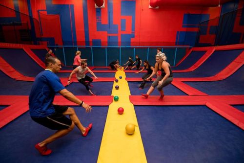 A vibrant indoor trampoline park with people playing dodgeball on trampolines, surrounded by colorful walls.