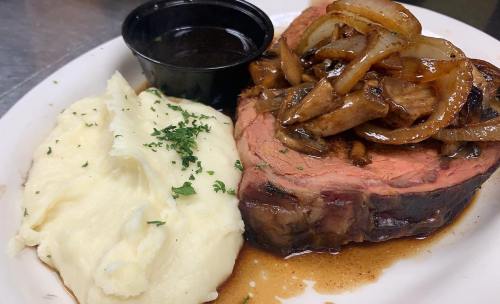 A plate featuring sliced roast beef topped with sautéed onions and mushrooms, served with mashed potatoes and gravy.