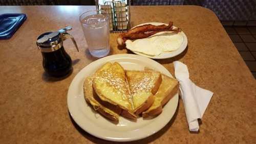 A plate with French toast, syrup, and a glass of water, alongside a plate of bacon and eggs.