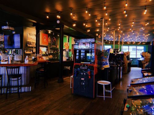 A cozy arcade bar with vintage game machines, a wooden bar, and warm string lights illuminating the space.