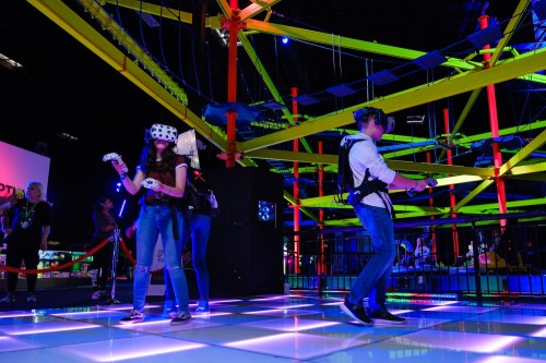 Two people wearing VR headsets and controllers stand on a brightly lit floor in a colorful, high-tech gaming environment.