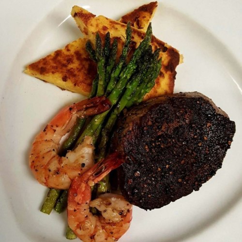 A plate featuring a grilled steak, shrimp, asparagus, and cornbread triangles, garnished and beautifully arranged.