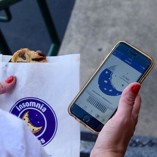 A person holds a cookie in a bag while checking a mobile app with a barcode for Insomnia Cookies.