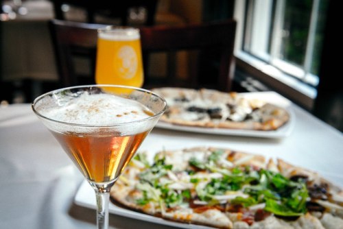A cocktail in a martini glass in the foreground, with a pizza and a beer in the background on a table.