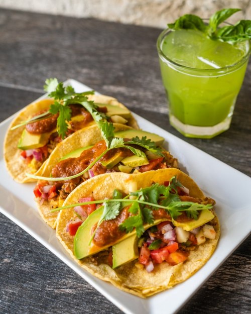 Three tacos topped with meat, avocado, and salsa, served on a white plate with a green drink in a glass.