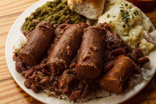 A plate of rice topped with sausage, beans, mashed potatoes, green peas, and a biscuit on the side.