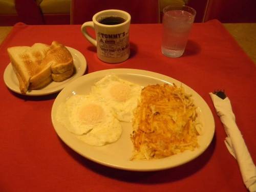 A plate with two fried eggs, hash browns, toast, a coffee mug, and a glass of water on a red tablecloth.