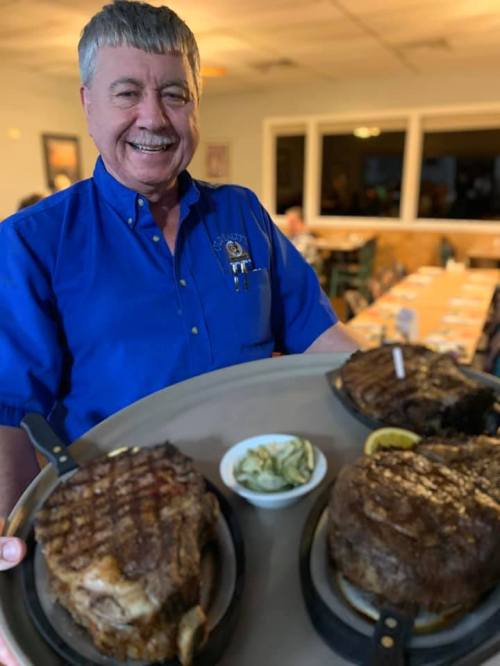 A smiling man in a blue shirt holds a tray with three grilled steaks and a small bowl of sauce.
