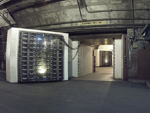 A dimly lit underground vault with large metal doors and exposed wiring, leading to another room.