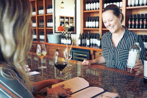 A smiling server presents a bottle of wine to a customer at a bar with wine bottles in the background.