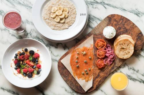 A marble table with a bowl of oatmeal, a fruit salad, smoked salmon on a wooden board, and drinks.