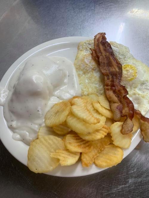 A plate of breakfast featuring eggs, bacon, biscuits with gravy, and crispy potato chips.