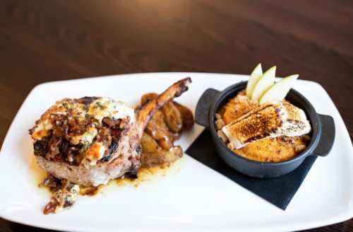 A plated meal featuring a grilled pork chop topped with cheese and bacon, served with roasted apples and a dessert.