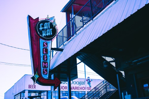 Neon sign for "Fat Kat Slams" with an arrow, set against a blue sky and nearby buildings.