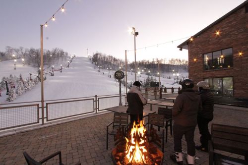 A cozy outdoor scene with a fire pit, overlooking a snowy ski slope and a rustic building at dusk.