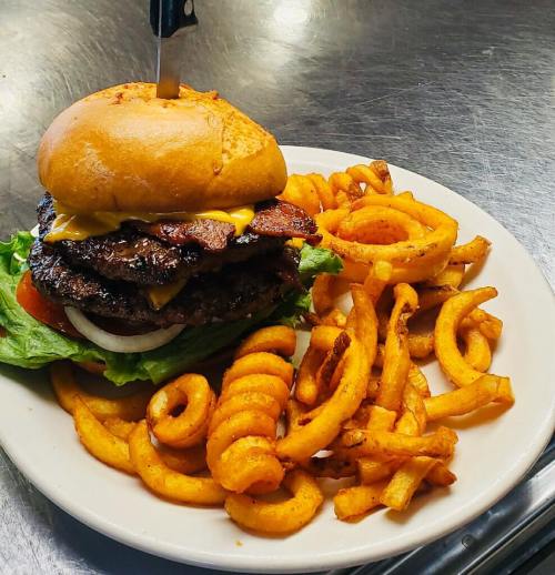 A stacked burger with cheese, bacon, lettuce, and tomato, served with curly fries on a white plate.