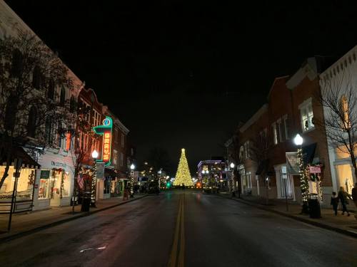 A festive street at night, featuring a large Christmas tree and decorated storefronts, creating a warm holiday atmosphere.