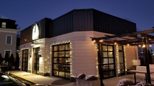 A modern restaurant exterior at dusk, adorned with string lights and large glass doors, surrounded by snow.