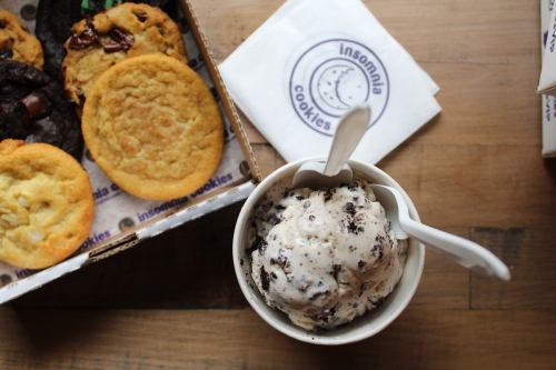 A bowl of cookies and cream ice cream with two spoons, next to a box of assorted cookies on a wooden table.