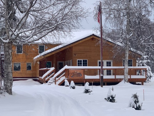 A wooden cabin surrounded by snow, featuring a front porch and an American flag, with trees in the foreground.