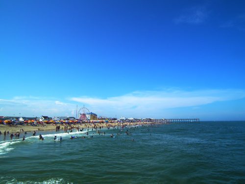 A sunny beach scene with people swimming and playing, a pier in the distance, and a ferris wheel on the shore.