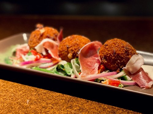 Three golden-brown croquettes on a bed of mixed greens, garnished with thin slices of prosciutto and colorful vegetables.