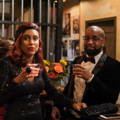A man and woman in formal attire hold drinks at a stylish event, with flowers and decor in the background.