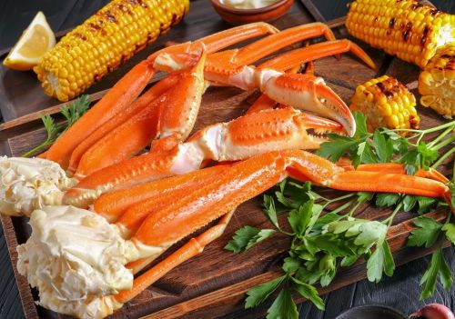 A wooden platter with cooked crab legs, grilled corn on the cob, and fresh parsley, garnished with lemon and sauce.