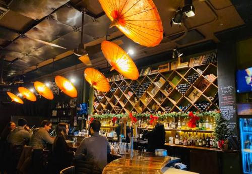 A cozy bar with orange umbrellas hanging above, shelves of wine, and patrons enjoying drinks in a warm atmosphere.