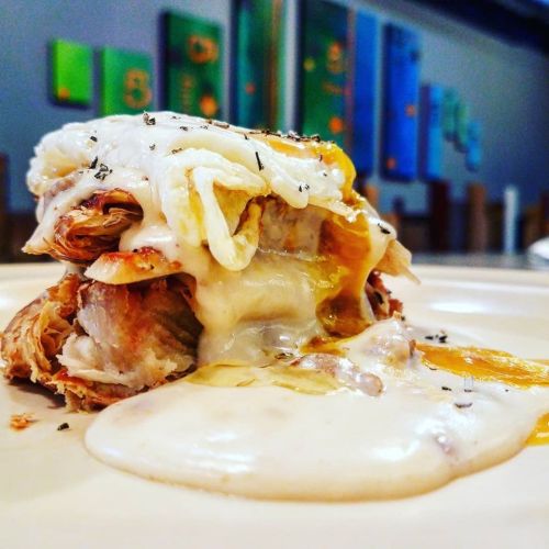 A close-up of a layered dish with melted cheese, a fried egg on top, and a creamy sauce, set against colorful artwork.