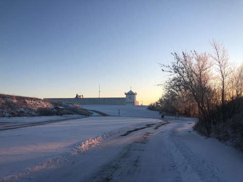 A snowy landscape with a road leading to a large building, surrounded by trees and a clear sky at sunrise.