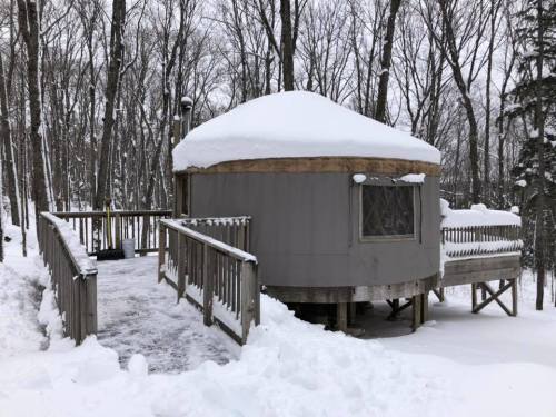 A cozy yurt covered in snow, surrounded by trees, with a wooden deck leading to the entrance.