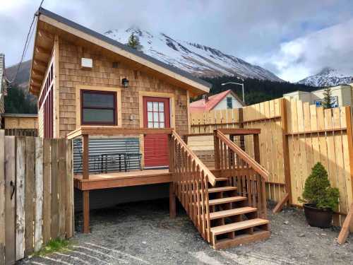 A small wooden house with a red door and stairs, surrounded by a wooden fence and mountains in the background.