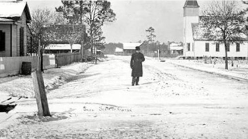A person walks down a snowy street lined with houses and a church in a historic black-and-white photograph.