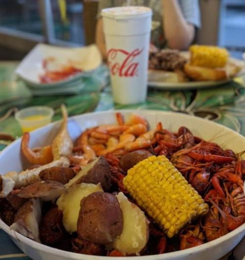 A large bowl of seafood with crawfish, shrimp, potatoes, and corn, with a soda cup in the background.