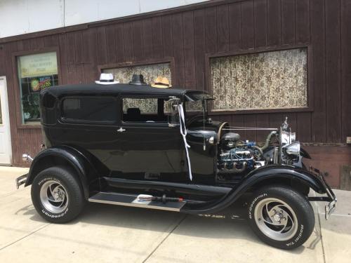 A vintage black car parked outside a building, adorned with hats on the roof and a shiny engine visible.