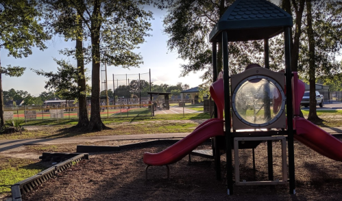Playground with a slide and climbing structure, surrounded by trees and a sports field in the background.