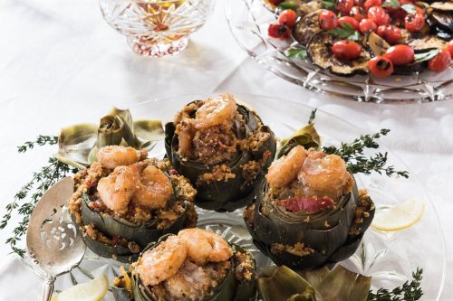 Three stuffed artichokes topped with shrimp, served on a white tablecloth with a side of roasted vegetables.