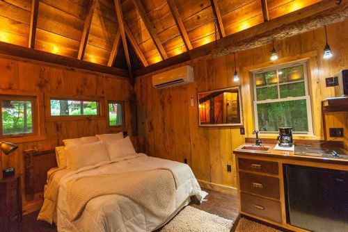 Cozy wooden cabin interior with a bed, kitchenette, and warm lighting, surrounded by nature.