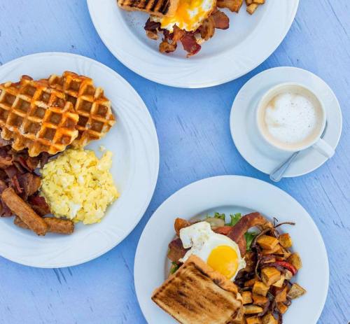 A breakfast spread featuring waffles, scrambled eggs, bacon, sausage, and a cup of coffee on a light blue table.