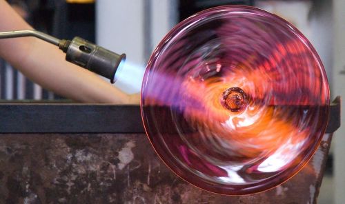 A glassblower shapes molten glass with a torch, creating a vibrant, swirling glass piece.