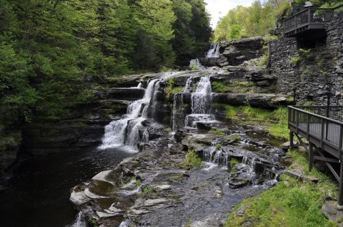 A serene waterfall cascades over rocky terrain, surrounded by lush greenery and a wooden viewing platform.