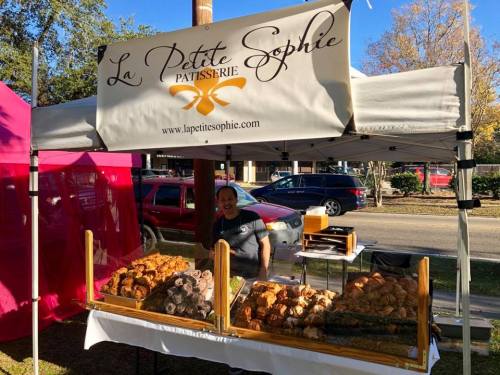 A smiling vendor stands behind a display of pastries at a market stall, with a sign for "La Petite Sophie Pâtisserie."