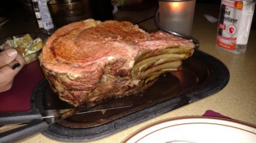 A large, juicy prime rib roast on a metal platter, with a knife and fork beside it, set on a restaurant table.