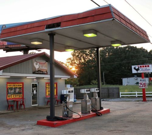 A gas station with a red roof, featuring a chicken restaurant, outdoor seating, and fuel pumps.